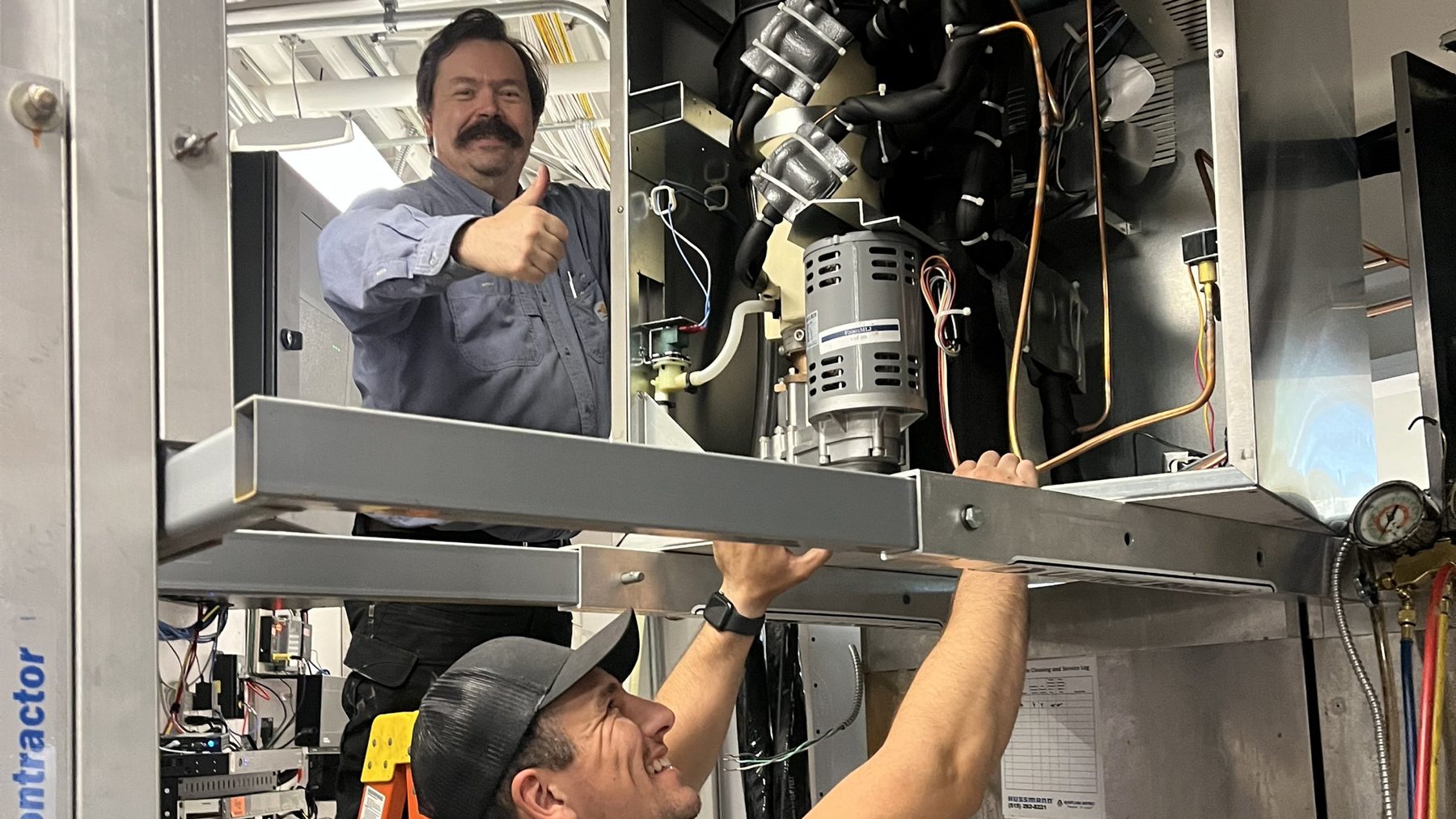 Two SRV technicians servicing an overhead refrigeration unit together, one working the panel and one giving a thumbs up, in Urbandale, Iowa